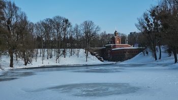 Картинки января / Парк Победы в Калининграде. Аусфальские ворота.
Аусфальские ворота, построенные в XIX веке располагались внутри земляного крепостного вала. Над проездом ворот находится боевая платформа, украшенная зубчатым парапетом. Ворота служили проходом через земляной вал и пропускали только пешеходов. С полевой стороны к воротам примыкал небольшой мост через крепостной ров, на месте которого теперь образовалось небольшое озеро. В XX в. единственный небольшой проезд заложили. Как выглядел фасад, обращенный в сторону города, остается загадкой, поскольку его засыпали землей. В 1993 г. на верхнем покрытии построили часовню Георгия Победоносца. Планируется реставрация ворот, и размещение в их помещениях музейных экспозиций. Январь 2026