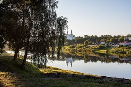 В лучах заходящего солнца. / Пронзают купола небесный свод, березки над рекой стоят склоненно. Река в своём величее течёт и все спокойно и умиротворённо .