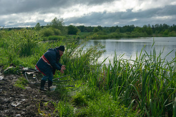 В ожидании клева / Рыбак подкармливает рыбу в озере,облачное небо,трава и лес